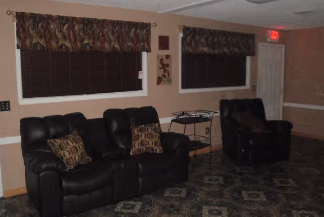 Dimly lit common room with leather sofa and recliner chairs against a wall beneath curtained windows.