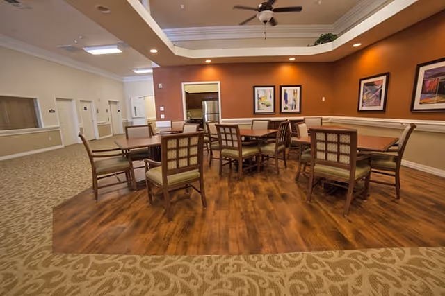 A dining area in an assisted living facility with several wooden tables and cushioned chairs arranged on a wood-finished floor section surrounded by carpet. The walls are painted in warm tones with framed artwork hanging, and there is a ceiling fan above.