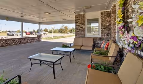 Covered outdoor patio area with cushioned seating including chairs and a sofa, two glass-top tables, a brick wall with a window, and a decorative floral wreath. The patio overlooks a parking lot with several cars and trees in the background.