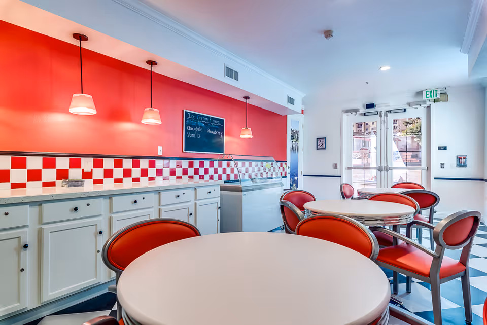 Retro-style dining room with round white tables, red vinyl chairs, a red-and-white checkered counter, pendant lights, and double exit doors.