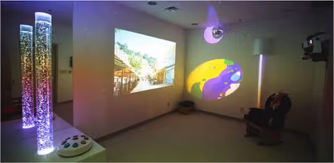 A dimly lit sensory room with bubble tubes emitting colorful light on the left, a projection of an outdoor scene on the wall, and a colorful abstract light projection on another wall. There is a comfortable chair in the corner and a small speaker or device on the floor.
