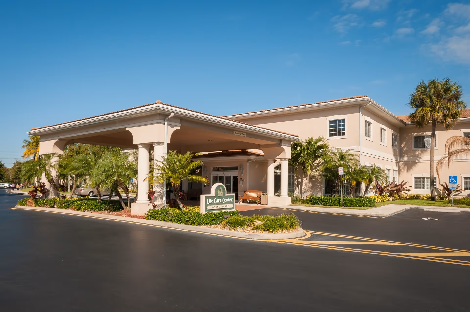 Front entrance and covered porte-cochere of the Life Care Center at Inverrary, a two-story beige building with palm trees and a driveway.