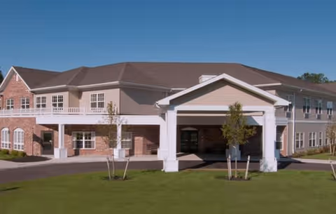 Front exterior of a two-story assisted living facility with a covered porte-cochere, brick and siding facade, and a manicured lawn.