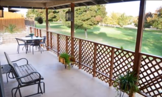 Covered porch with benches, a table, and potted plants overlooking a grassy yard and trees.