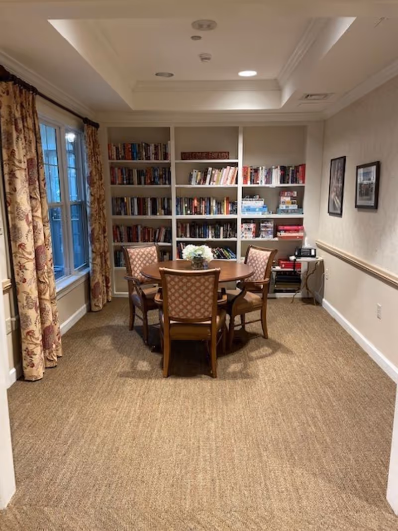 A cozy room with a round wooden table surrounded by four upholstered chairs. Behind the table is a built-in bookshelf filled with books and board games. The room has beige carpet, floral curtains on the window to the left, and two framed pictures on the right wall.