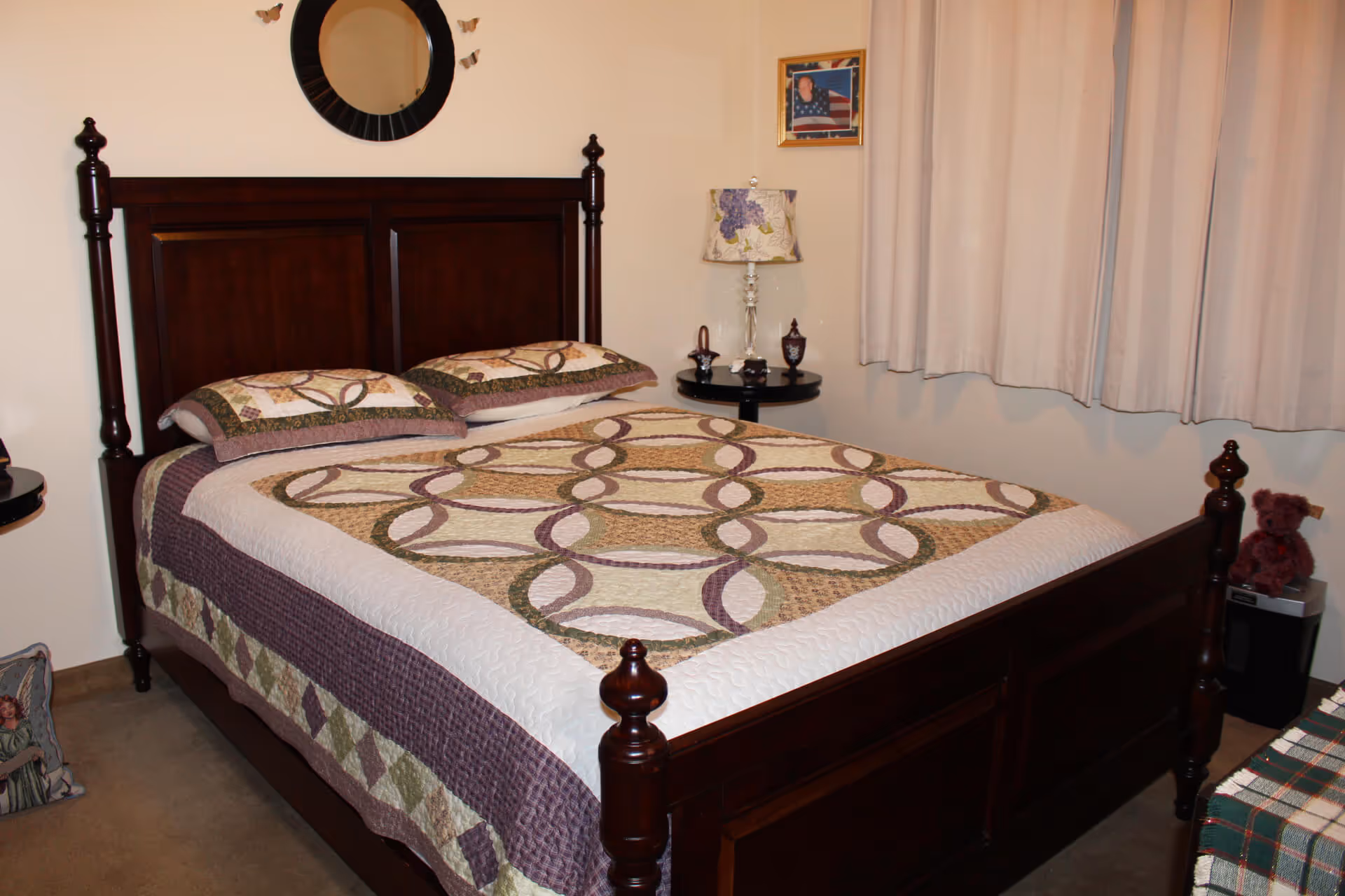A bedroom with a dark wooden bed frame and a quilted bedspread featuring a circular pattern in shades of beige, green, and purple. Two matching pillows are on the bed. A round mirror and small butterfly decorations hang on the wall above the headboard. There is a small round side table with a floral lampshade, a few decorative items, and a framed photo on the wall. Beige curtains cover the window, and a teddy bear sits on a small black table in the corner.