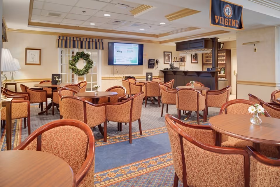 A cozy dining area with multiple round wooden tables surrounded by upholstered chairs with a red and gold pattern. A small bar area is visible in the background with bottles and glasses. A flat-screen TV is mounted on the wall above the bar, and a wreath hangs on a glass door. The room has beige walls, a patterned carpet, and a Virginia flag hanging from the ceiling.