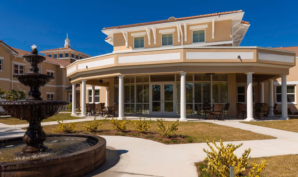 Exterior view of HarborChase of Villages Crossing showing a beige building with white columns supporting a covered patio area with tables and chairs. In the foreground, there is a multi-tiered water fountain surrounded by a circular stone basin, landscaped with small bushes and grass under a clear blue sky.