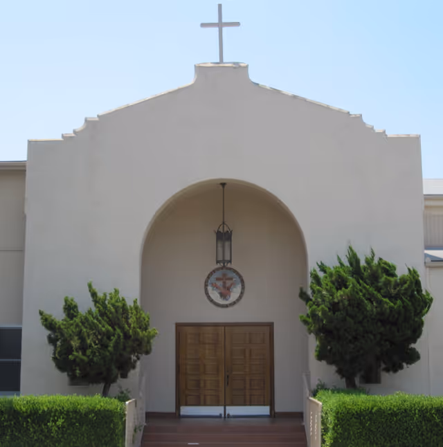 Front exterior view of a building with a white facade, a cross on top, a large arched entrance with a hanging lantern, a circular emblem above double wooden doors, and green bushes on either side of the entrance.