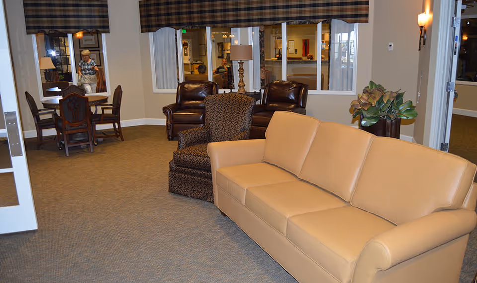 Interior view of a senior living facility common area with a beige leather sofa, a patterned armchair, two dark brown leather chairs, a wooden table with four chairs, a lamp on a side table, and a large plant. There are windows and a door visible in the background, with a person walking in the distance.