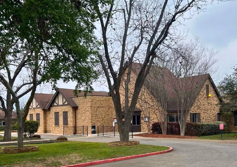 Exterior view of a brick building with a brown roof, surrounded by leafless and leafy trees, a driveway, and a grassy area with a red curb.