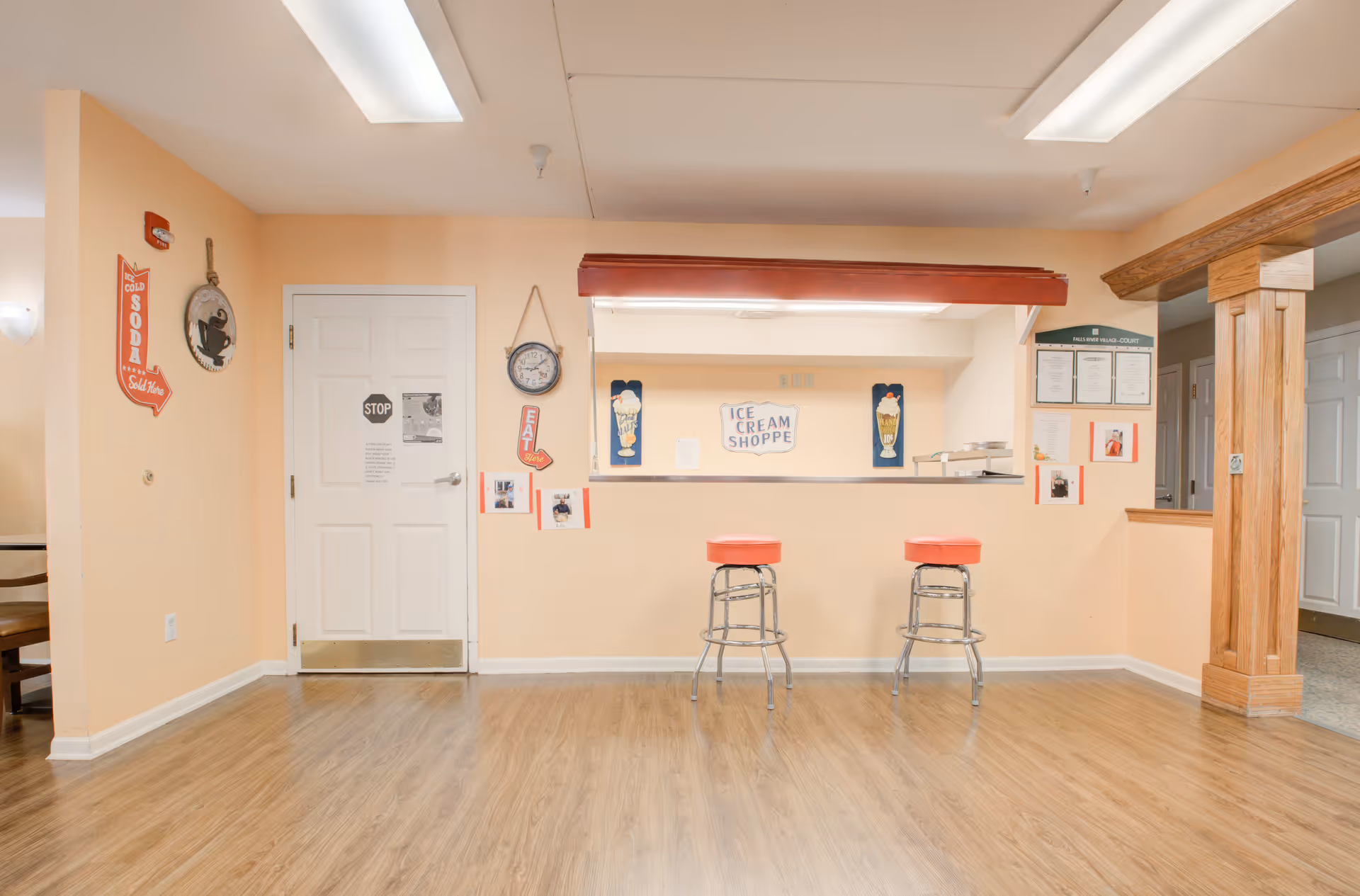 Interior view of a memory care facility room with light peach walls and wood flooring. There is a counter with two red cushioned bar stools in front of it. Above the counter is a sign that reads 'Ice Cream Shoppe' with pictures of ice cream sundaes on either side. On the left wall, there are vintage-style signs including one that says 'Cold Soda Sold Here' and a clock. To the right, wooden columns frame a hallway with white doors.