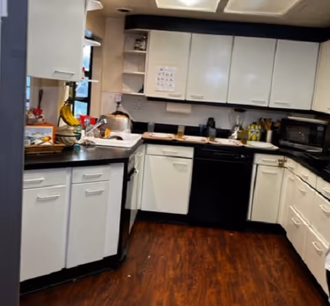 Interior view of a kitchen with white cabinets, black countertops, and wooden flooring. Various kitchen appliances and items such as a microwave, blender, bananas, and a toaster are visible on the counters.