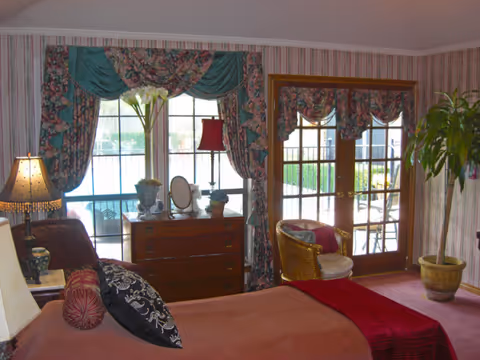 Cozy bedroom with pink bedspread, floral drapes, dresser, and French doors.