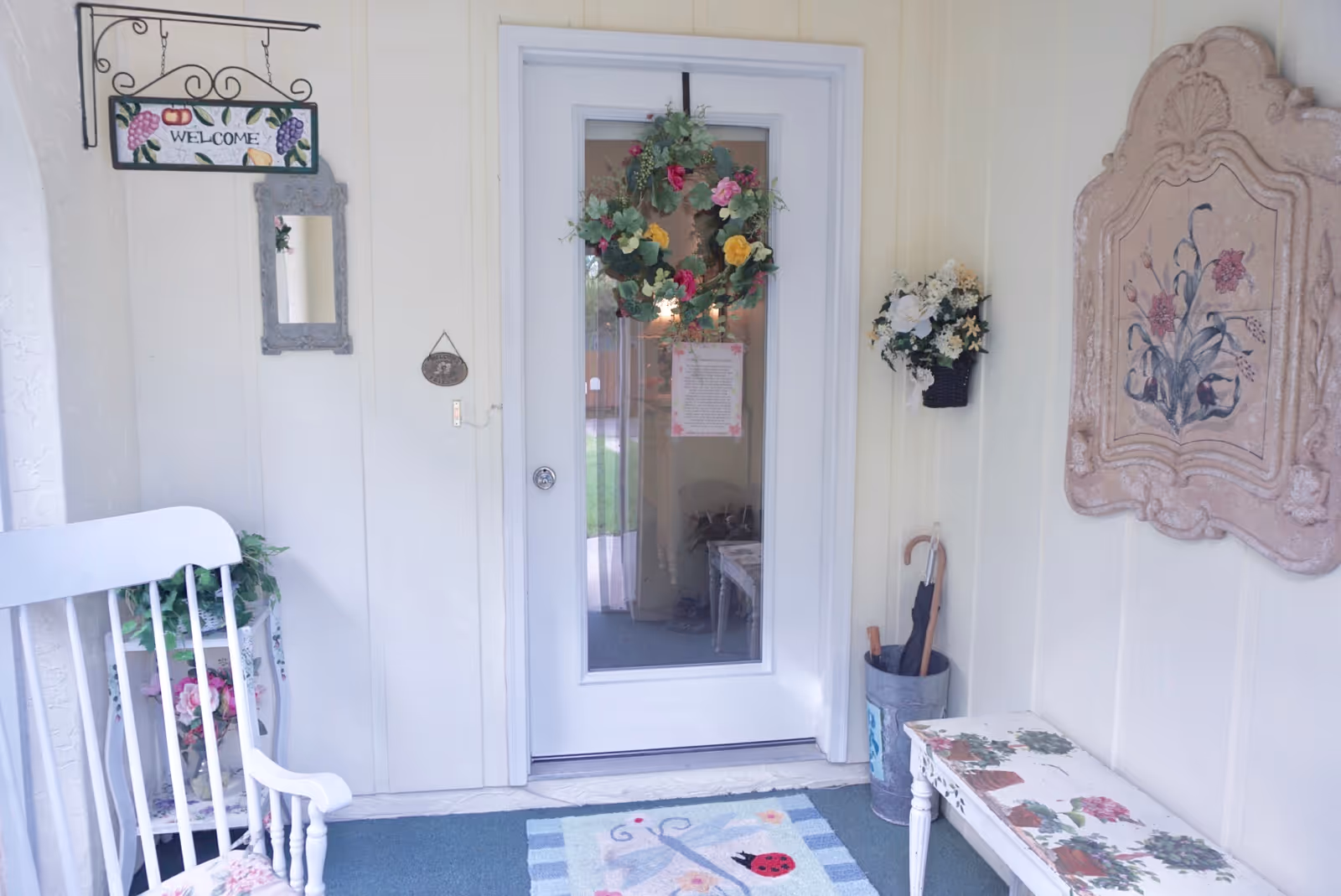 A cozy entrance area with a white door decorated with a floral wreath. To the left, there is a white rocking chair and a small table with a plant. A welcome sign hangs on the wall above a small mirror. To the right, there is a floral wall decoration, a small bench with floral patterns, and an umbrella stand with umbrellas. A colorful rug with a ladybug design is placed in front of the door.