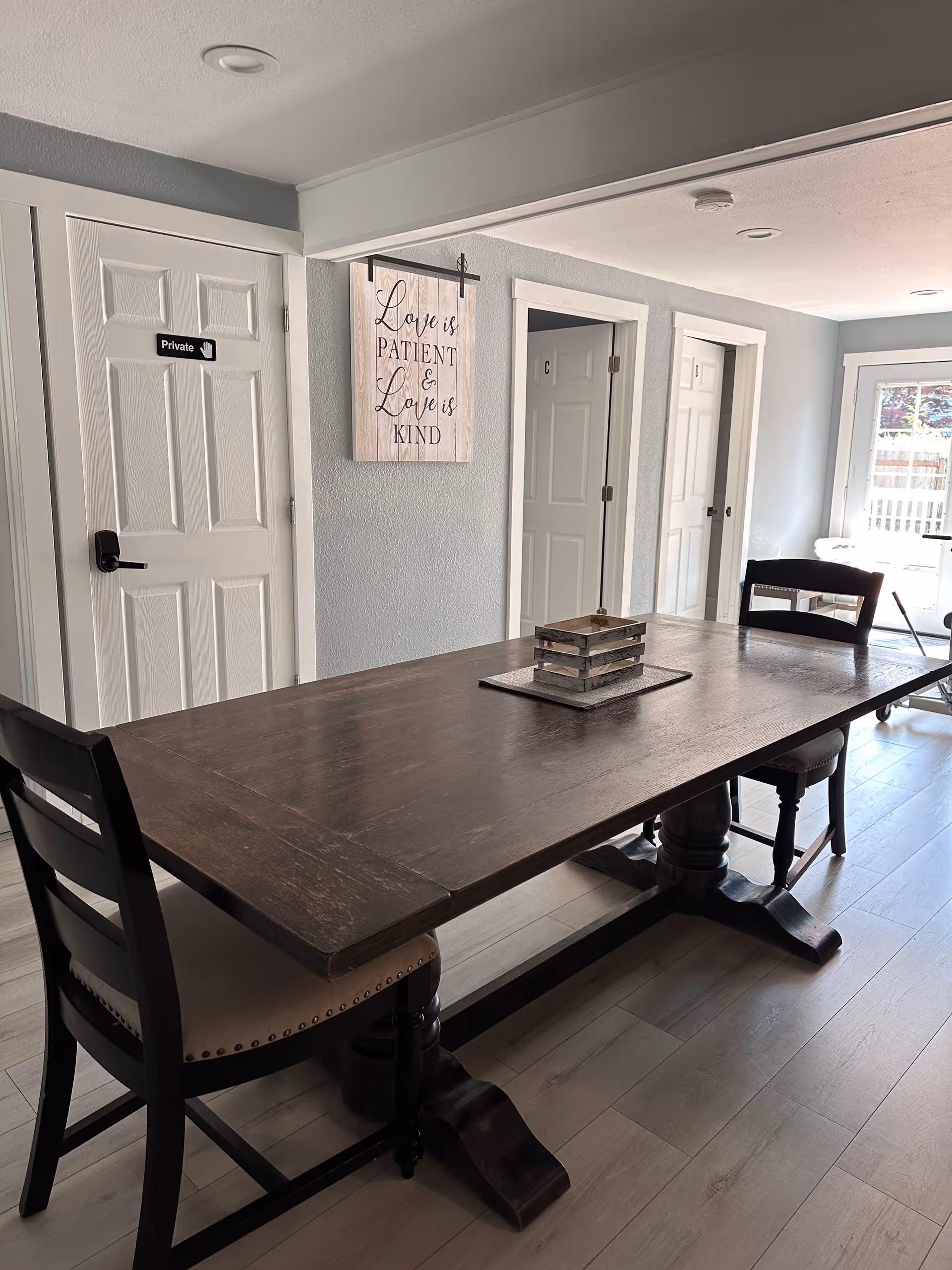 A dining area with a long dark wooden table and two chairs on either side. On the wall behind the table, there is a decorative sign that reads 'Love is patient & Love is kind.' There are three white doors along the wall, one labeled 'Private.' The room has light-colored walls and flooring, with a door leading outside visible in the background.