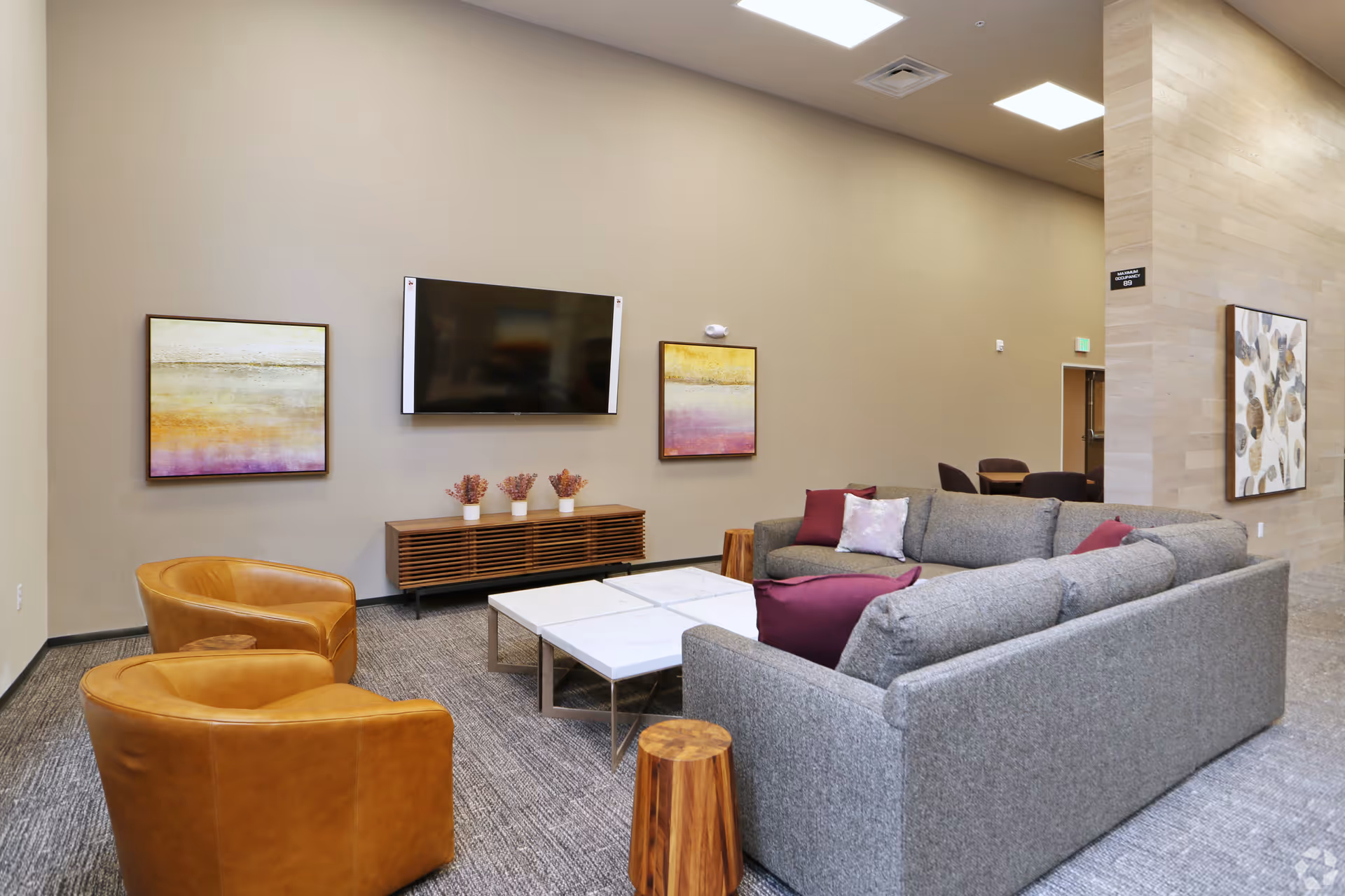 A modern living room area with a large gray sectional sofa adorned with purple and white pillows, two tan leather armchairs, and a set of four white marble coffee tables with metal legs. The room features a wall-mounted flat screen TV above a wooden media console with three small potted plants. The walls are decorated with abstract paintings in shades of yellow, purple, and brown. The space has a carpeted floor and a high ceiling with recessed lighting.