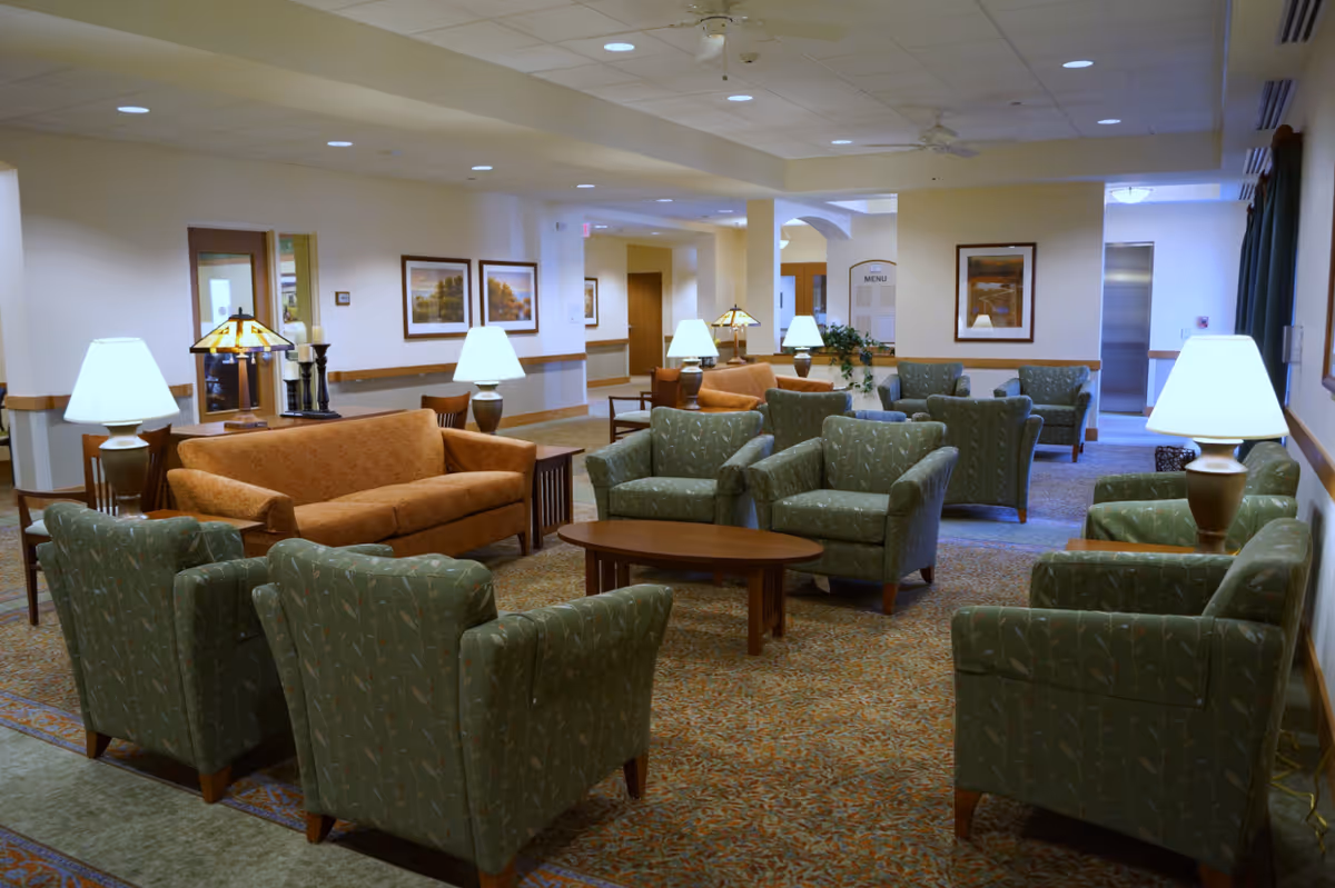 Spacious communal lounge with multiple green armchairs and orange sofas arranged around coffee tables and lamps.