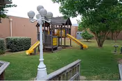 Outdoor playground area with a wooden play structure featuring two yellow slides, surrounded by green grass, trees, and a brick wall. A white lamp post with multiple globes is visible in the foreground.
