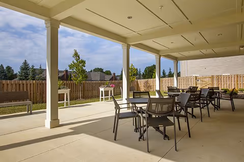 Covered outdoor patio with multiple tables and chairs, a fenced yard, and houses in the background.