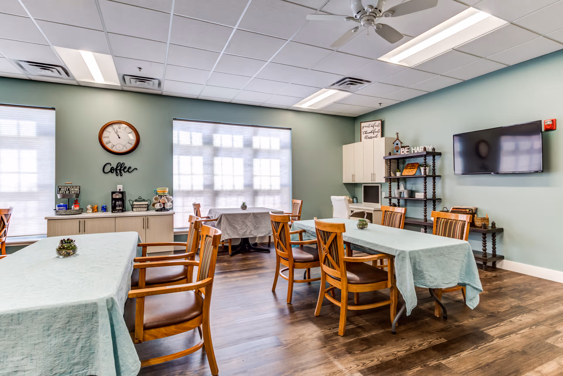 A bright room with light green walls and wooden flooring featuring several tables covered with light blue tablecloths and surrounded by wooden chairs with brown cushions. On the left wall, there is a coffee station with a clock above it and the word 'Coffee' displayed. The room also has a wall-mounted TV, a shelving unit with decorative items, and a small desk with a white chair. Large windows with blinds allow natural light to fill the space.