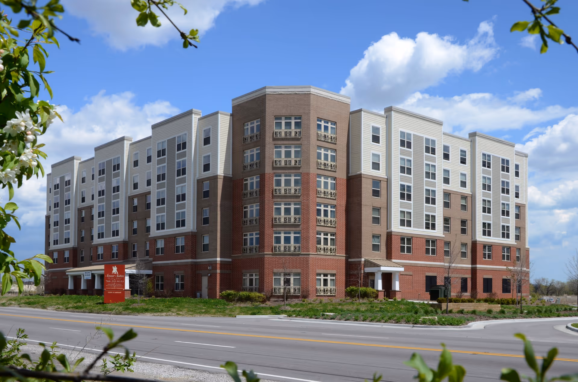 Exterior view of a multi-story senior living facility building with a mix of brick and light-colored siding under a partly cloudy blue sky. The building has many windows and a red sign near the entrance that reads Senior Suites of Fay’s Point.