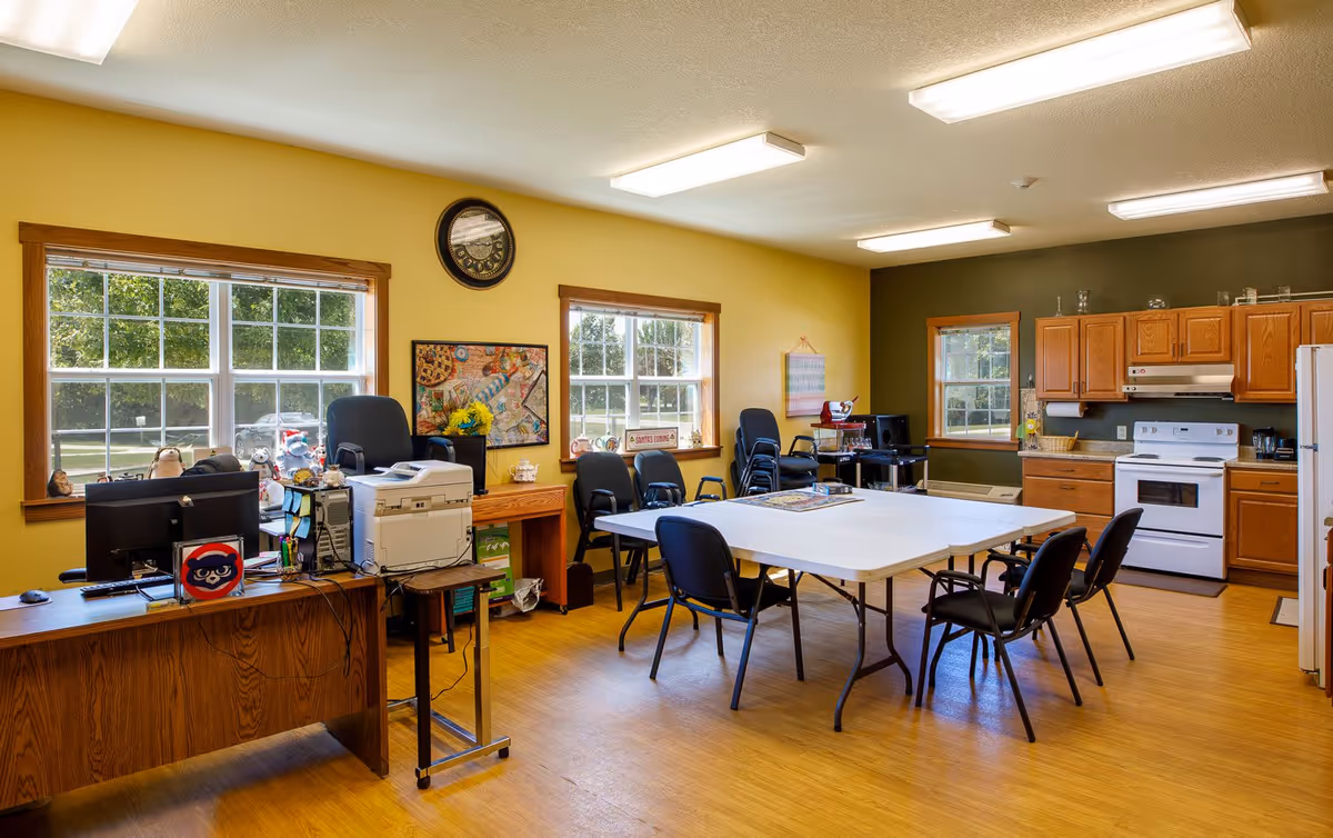 A bright room with yellow and green walls featuring a kitchen area with wooden cabinets, a white stove, and a refrigerator. In the center, there are two white folding tables pushed together surrounded by black chairs. On the left side, there is a wooden desk with computer monitors, a printer, and various office supplies. Large windows let in natural light and show green trees outside. The room has a wooden floor and fluorescent ceiling lights.