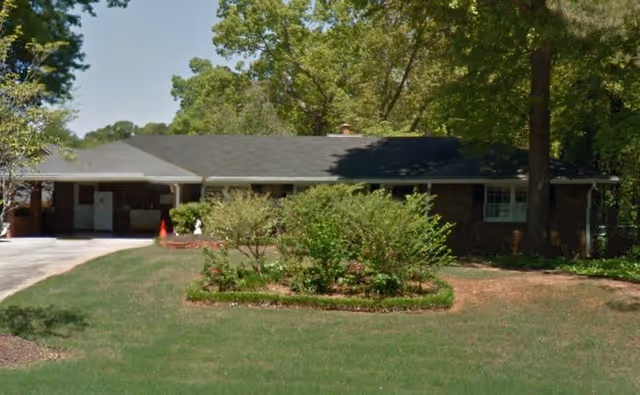 Single-story brick building with a dark roof surrounded by trees and bushes, with a driveway on the left side and a well-maintained lawn in front.