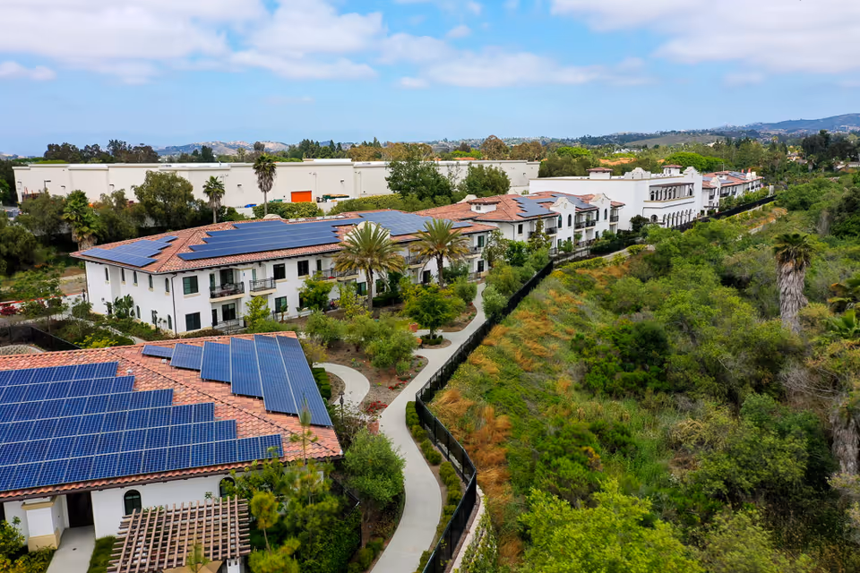 Aerial view of Oceanside Senior Living facility showing multiple buildings with red tile roofs equipped with solar panels. The buildings are surrounded by landscaped gardens, palm trees, and a winding pathway. There is a green, wooded area adjacent to the facility and a partly cloudy sky overhead.