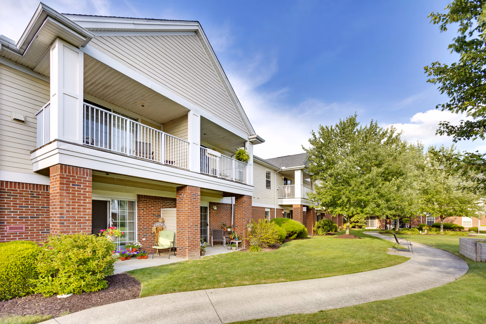 Exterior view of a senior living facility building with a covered balcony and patio area. The building features brick and light-colored siding, surrounded by well-maintained green lawns, shrubs, and trees under a blue sky with some clouds. A curved concrete walkway leads through the landscaped grounds with benches along the path.