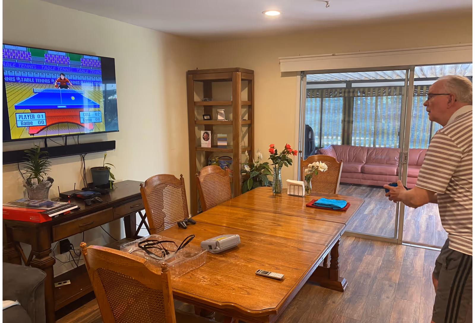 An elderly man playing a video game on a wall-mounted TV in a room with a wooden dining table and chairs. The TV screen shows a table tennis game. The room has wooden flooring, a wooden shelf with decorative items, and sliding glass doors leading to a sunroom with a pink couch.