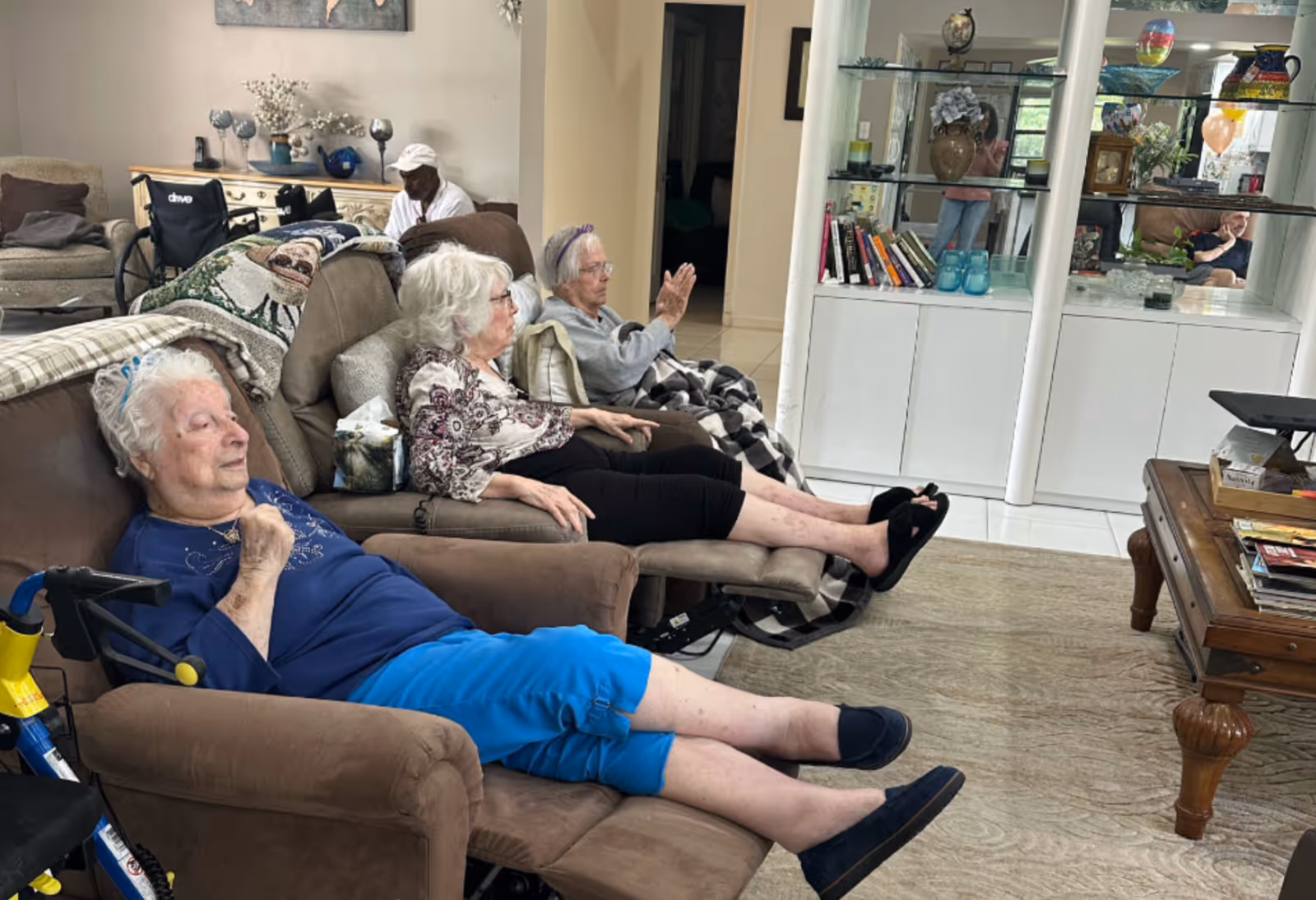 Three elderly women reclining in armchairs in a communal living room with shelving and a coffee table nearby.