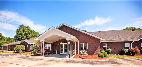 Exterior view of a single-story brick building with a covered entrance and a driveway in front, surrounded by small bushes and trees under a blue sky with some clouds.