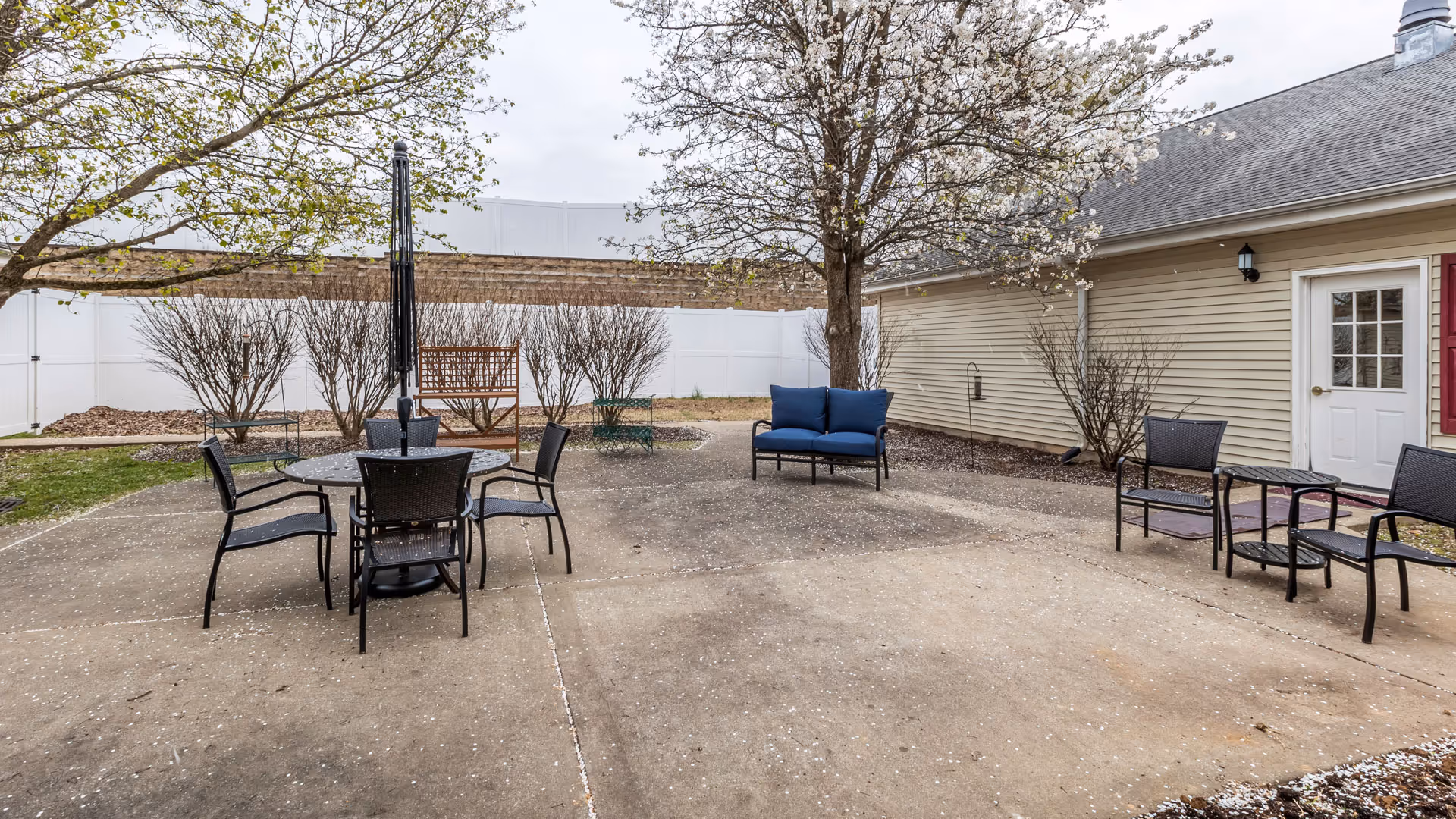 Outdoor patio area with several black metal chairs and a round table with an umbrella stand in the center. There is a blue cushioned bench and additional chairs arranged around the concrete patio. Trees with sparse leaves and a beige building with a white door are visible in the background.