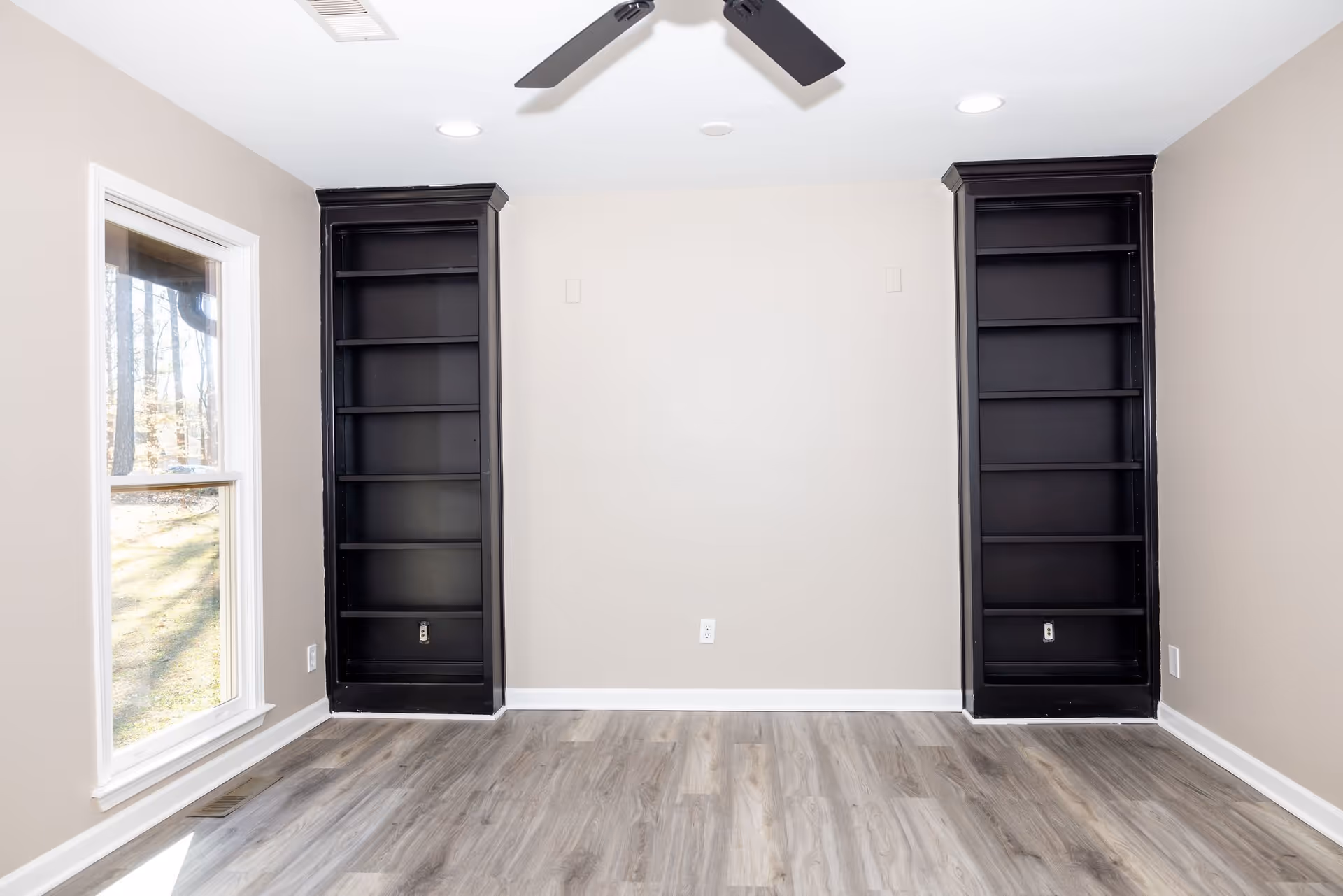 Empty living room with gray wood floors, two tall black built-in bookshelves flanking a blank beige wall, a ceiling fan, and a window on the left.