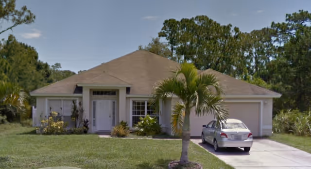 Single-story residential house with a beige roof, white exterior walls, a front door with sidelights, two windows, a two-car garage, a driveway with a parked silver car, a small palm tree in the front yard, and surrounding greenery including trees and bushes.