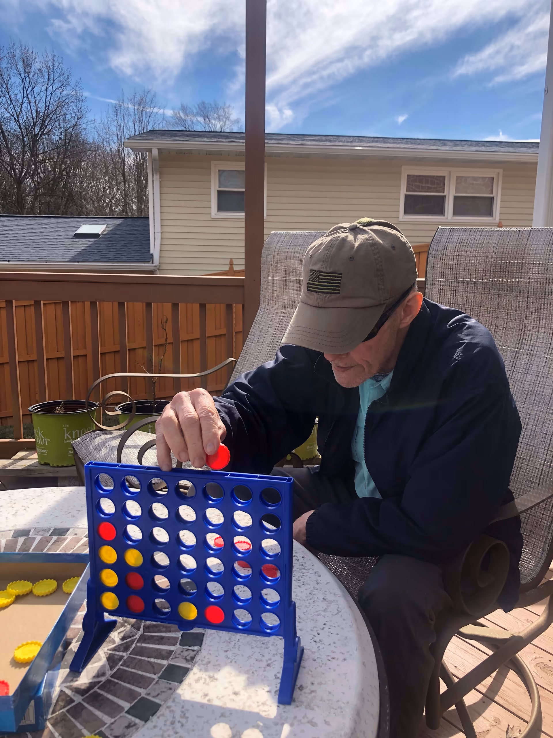 An elderly man wearing a cap and jacket is sitting outside on a patio playing a game of Connect Four on a round table. The background shows a wooden fence, some potted plants, and a house with beige siding under a partly cloudy sky.