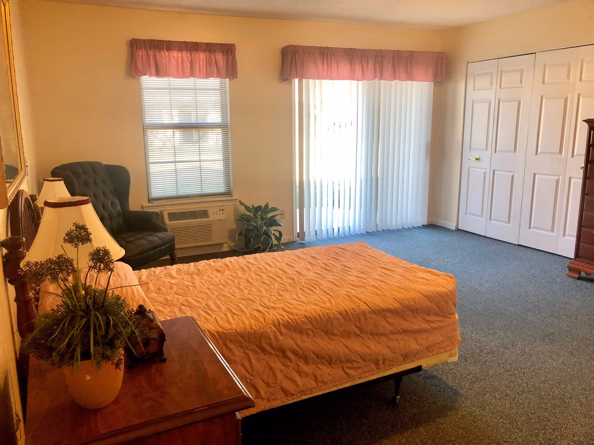 A bedroom with a bed covered in a peach quilt, a wooden nightstand with a lamp and a potted plant, a green upholstered armchair near a window with blinds, a sliding glass door with vertical blinds, and a double-door closet.