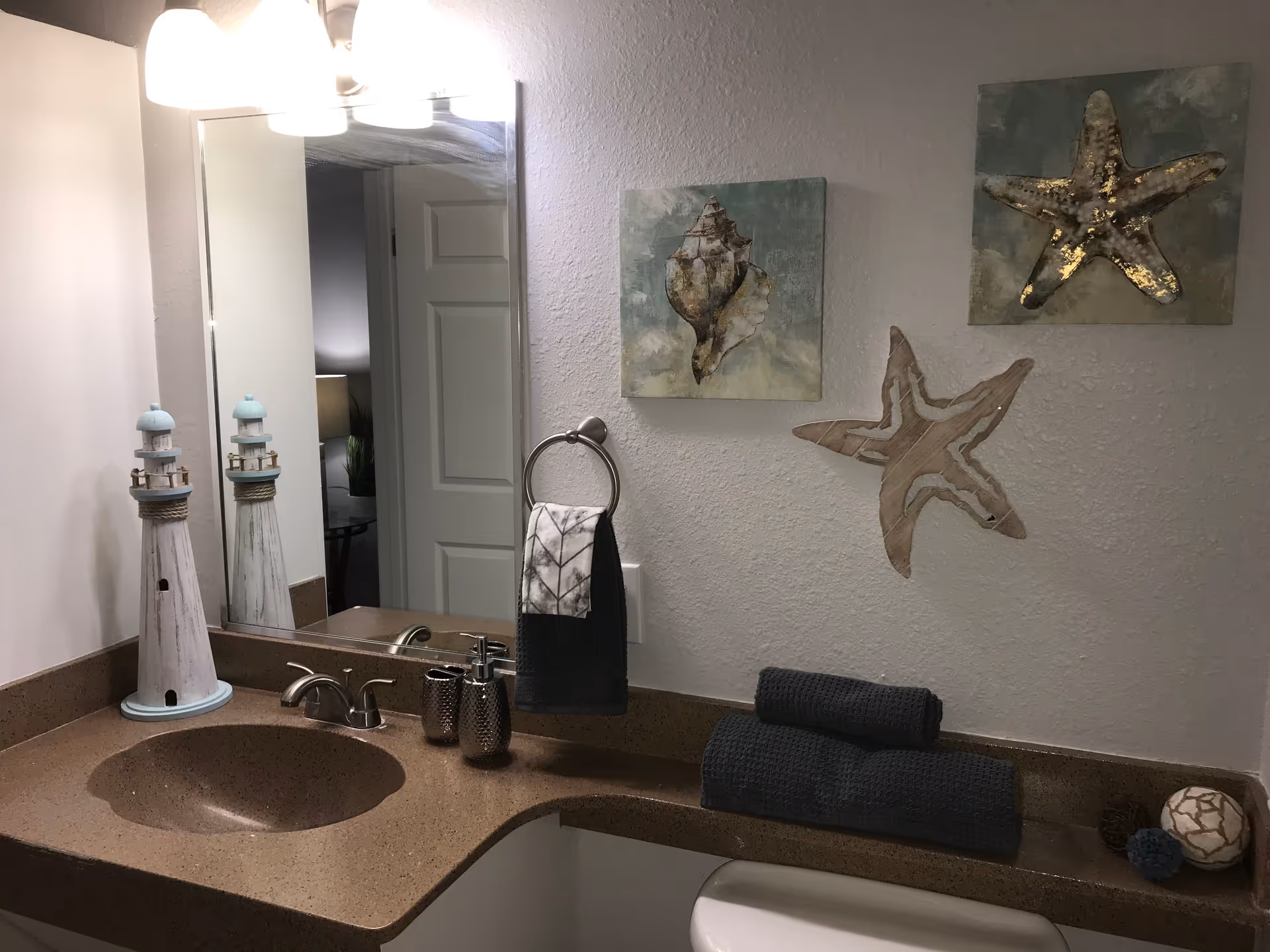 Bathroom sink area with a brown countertop, a silver faucet, a large mirror, and wall decorations including two framed pictures of sea shells and a wooden starfish. There are two rolled dark towels on the counter, a towel ring with a hand towel, and a decorative lighthouse figurine.