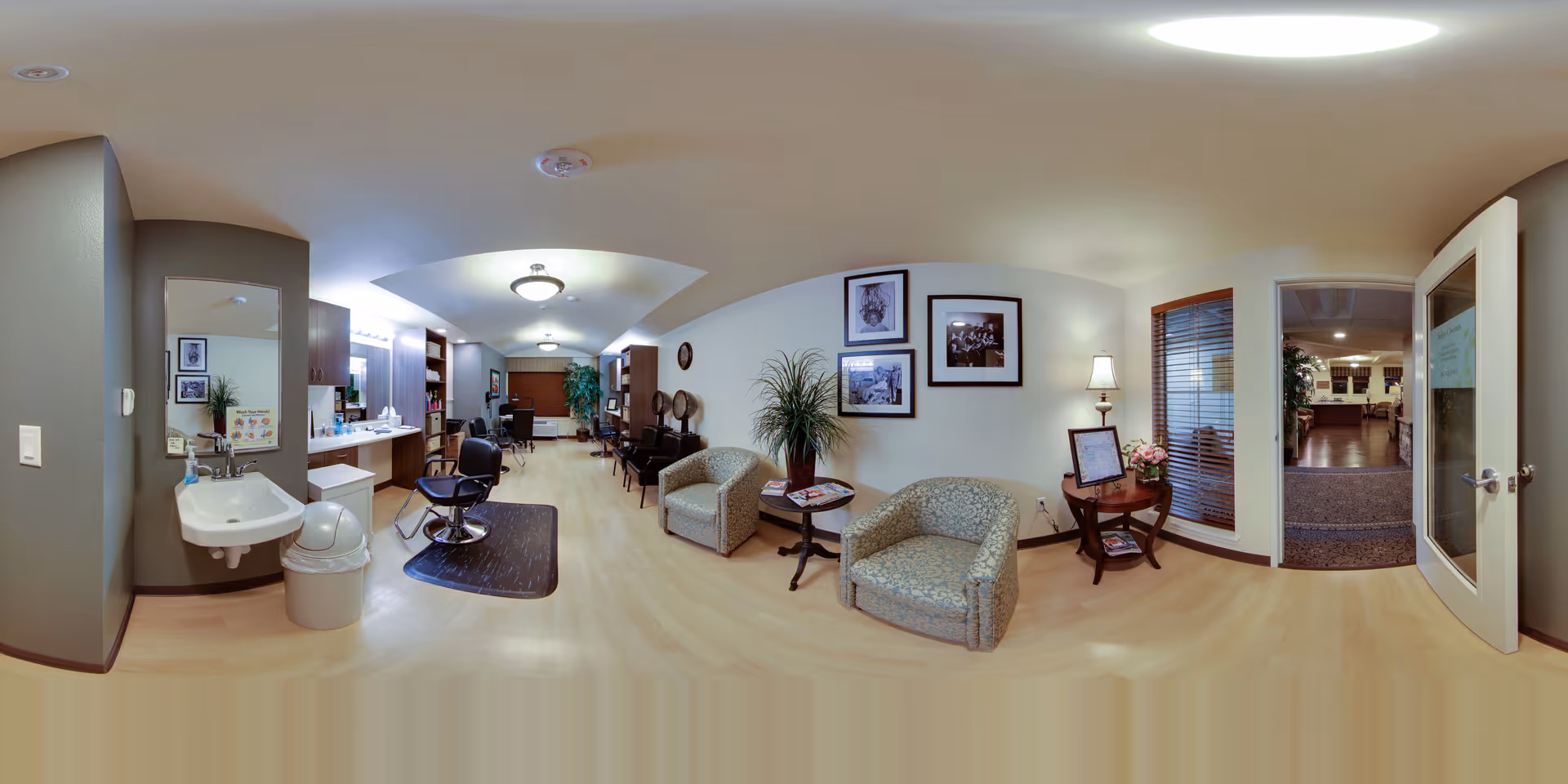 Spacious senior living facility interior showing a salon area with styling chairs and sink on the left, a seating/waiting area with armchairs and tables in the center, and an open doorway to another common area on the right.