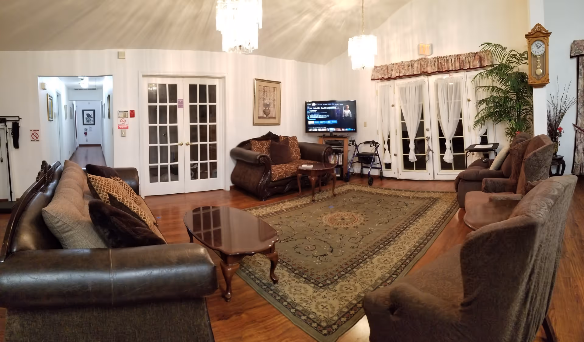 A spacious living room with multiple upholstered sofas and armchairs arranged around a large patterned area rug. There are two wooden coffee tables, a wall-mounted flat-screen TV showing a movie selection screen, a walker near the TV stand, and a grandfather clock on the wall. French doors with sheer curtains lead outside, and the room is illuminated by two hanging chandeliers. The floor is wooden, and there is a hallway visible to the left.