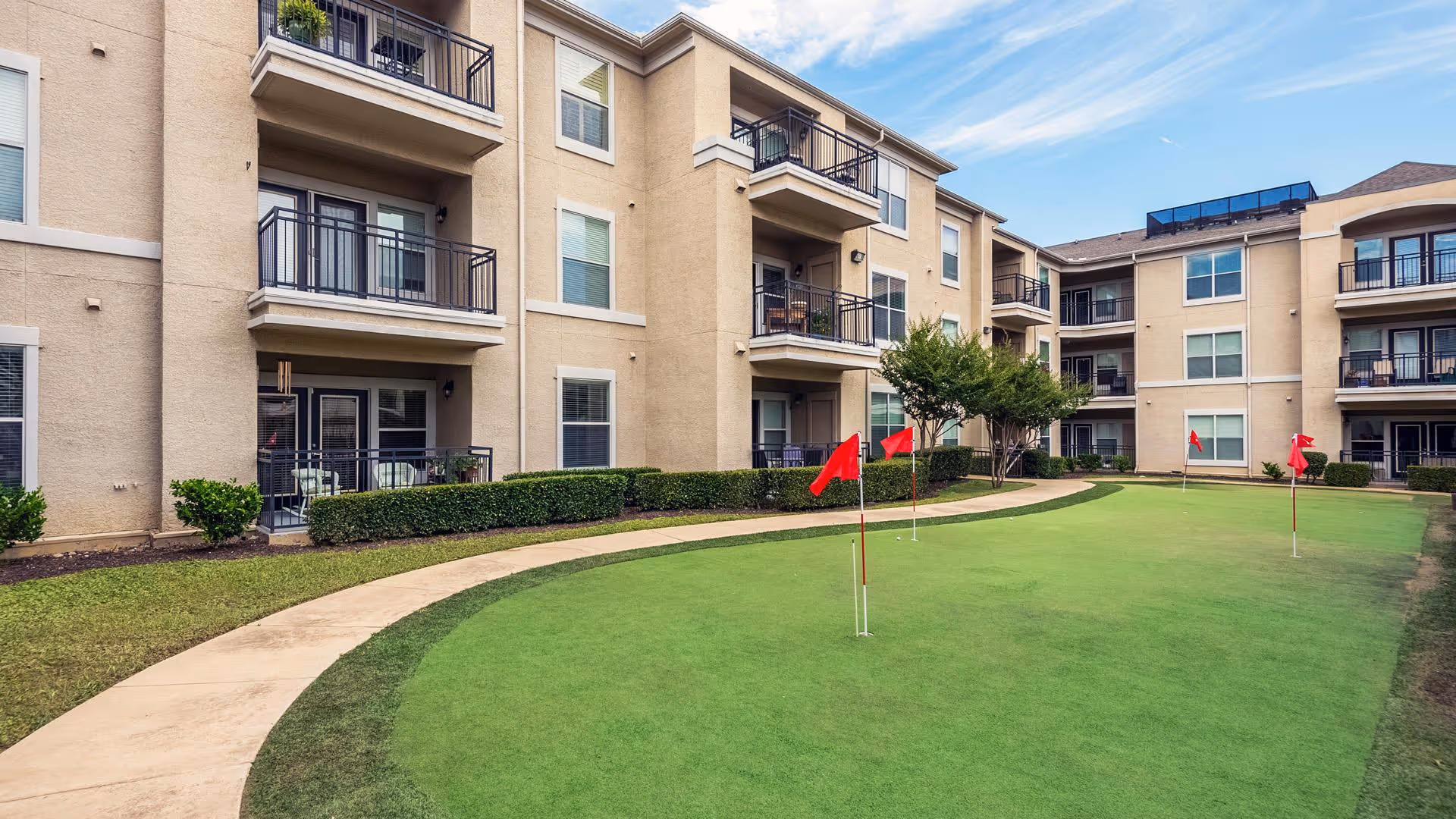 Outdoor courtyard area of a senior living facility with a putting green featuring several red flags. Surrounding the green are beige multi-story buildings with balconies and windows. A curved concrete walkway borders the putting green, and there are some bushes and small trees around the area under a partly cloudy sky.
