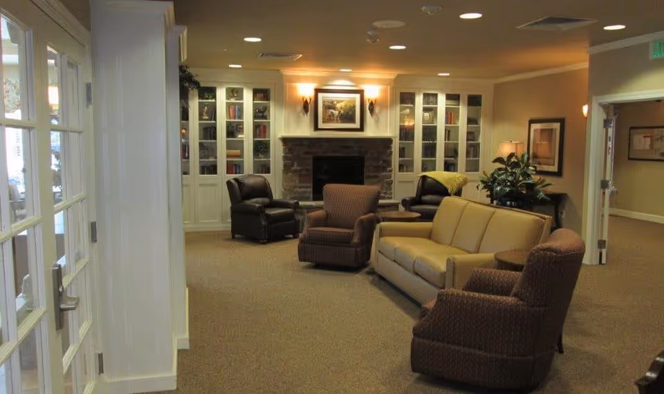 A cozy living room area in a senior living facility featuring a beige sofa, two patterned armchairs, and two dark leather armchairs arranged around a stone fireplace with built-in white bookshelves on either side. The room is softly lit with wall sconces and a table lamp, and there is a plant on a side table near the sofa. The walls are painted a warm beige color, and there is a framed picture above the fireplace.