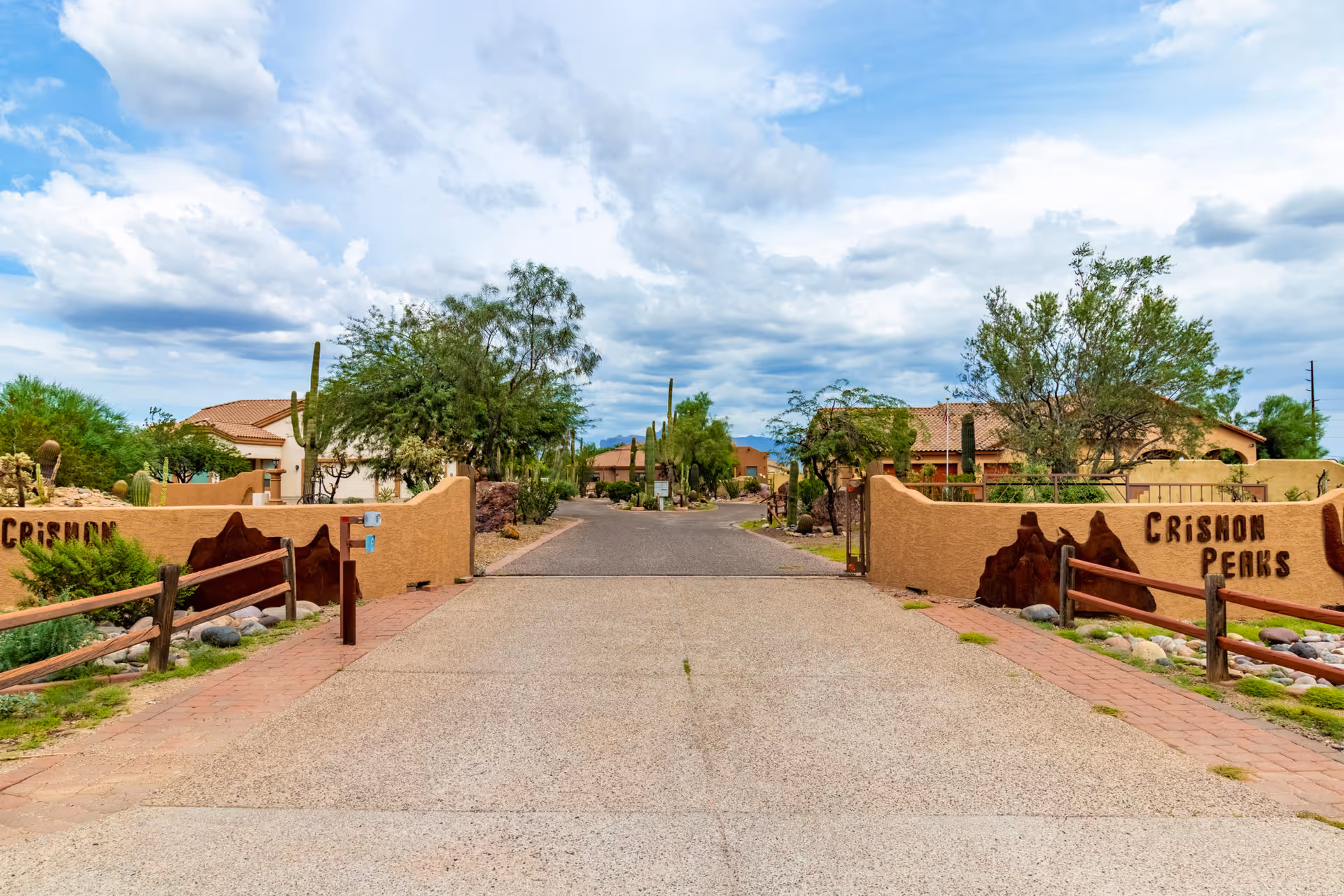 Entrance driveway to Crismon Peaks Assisted Living facility with desert landscaping, trees, and houses visible under a partly cloudy sky.