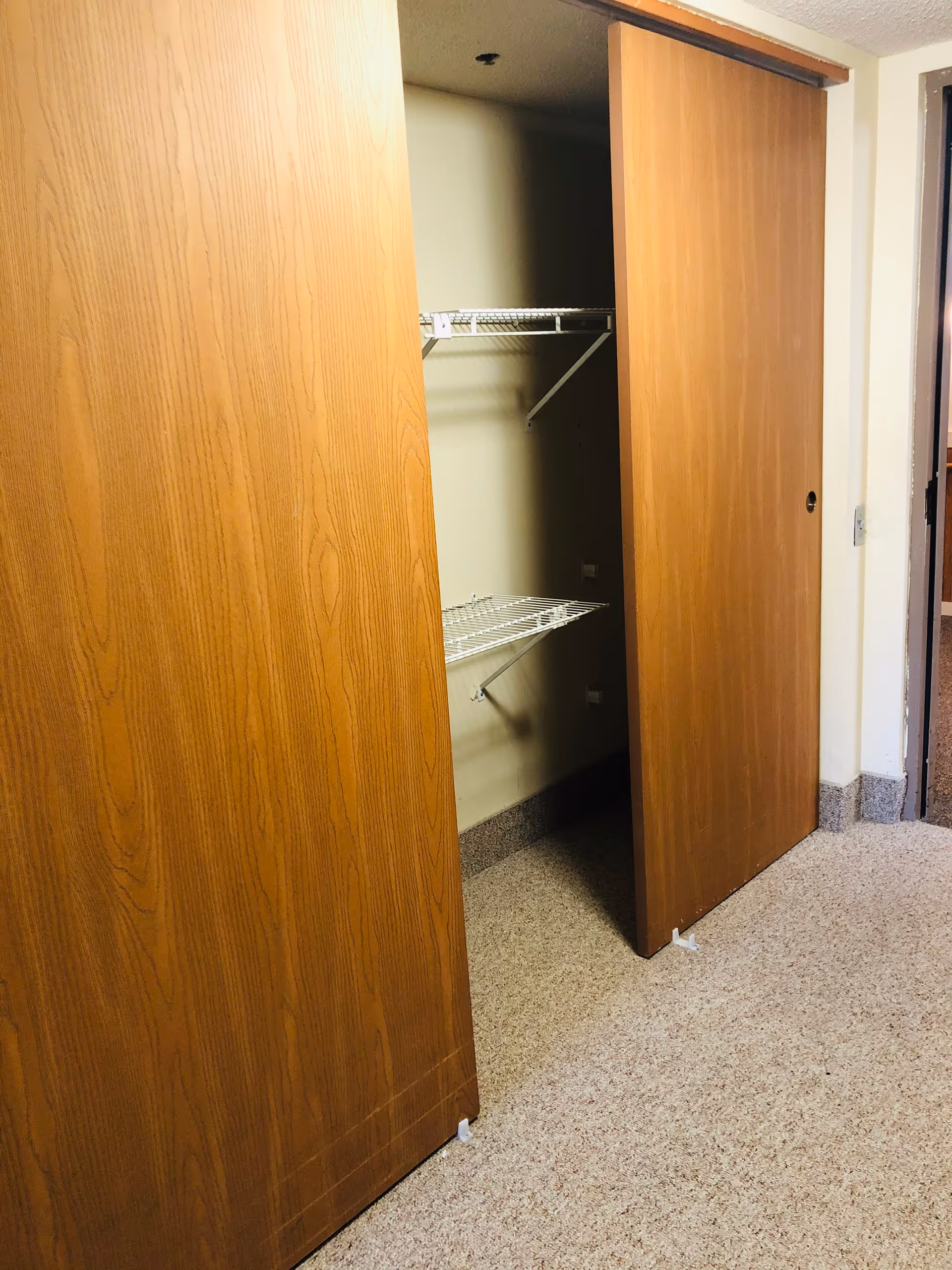 An open closet with two wooden sliding doors, revealing wire shelving inside. The floor is carpeted and the walls are light-colored.