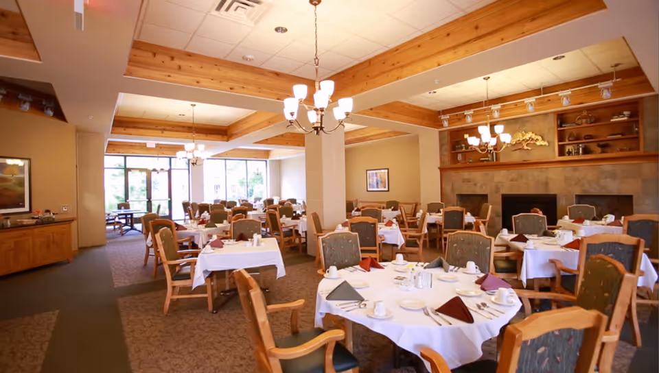 A spacious dining room in Tudor Oaks Senior Living Community with multiple round and square tables covered with white tablecloths, set with plates, cups, and folded napkins. The room features wooden beams on the ceiling, chandeliers, carpeted floors, and large windows letting in natural light. There is a fireplace with shelves above it on one side of the room.