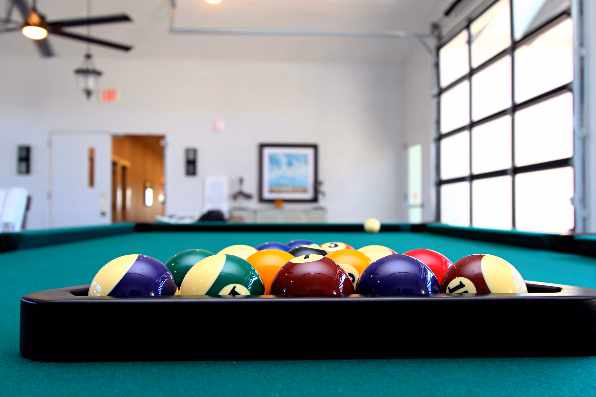 Racked billiard balls on a pool table in a bright recreation room with large windows and ceiling fans.