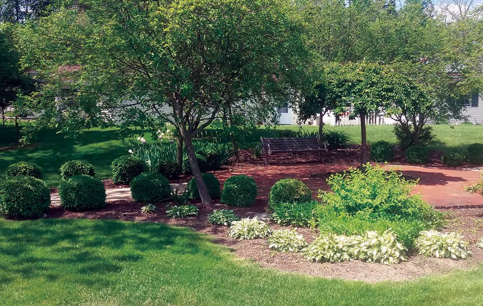 A well-maintained garden area with neatly trimmed bushes, green grass, and several trees providing shade. There is a wooden bench on a brick-paved area surrounded by plants and shrubs, with a building partially visible in the background.