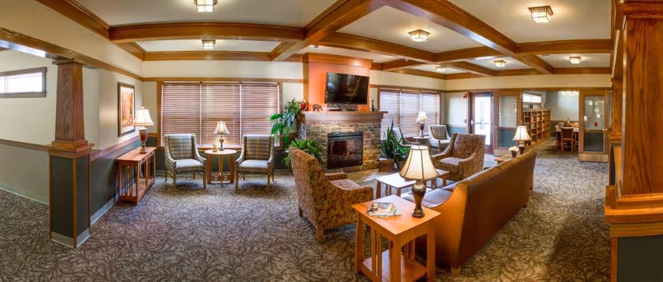 A spacious and warmly decorated living room in an assisted living facility featuring a stone fireplace with a mounted TV above it, multiple armchairs and sofas arranged around coffee tables, several table lamps providing soft lighting, large windows with blinds, wooden ceiling beams, and a carpeted floor with a floral pattern. There is also a small dining or activity area visible in the background.