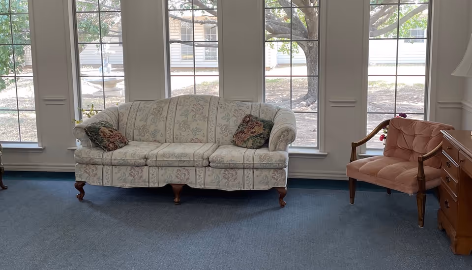 A living room area with a floral patterned sofa with two decorative pillows, a pink cushioned armchair, a wooden side table with drawers, and large windows showing an outdoor view with trees and a fountain.